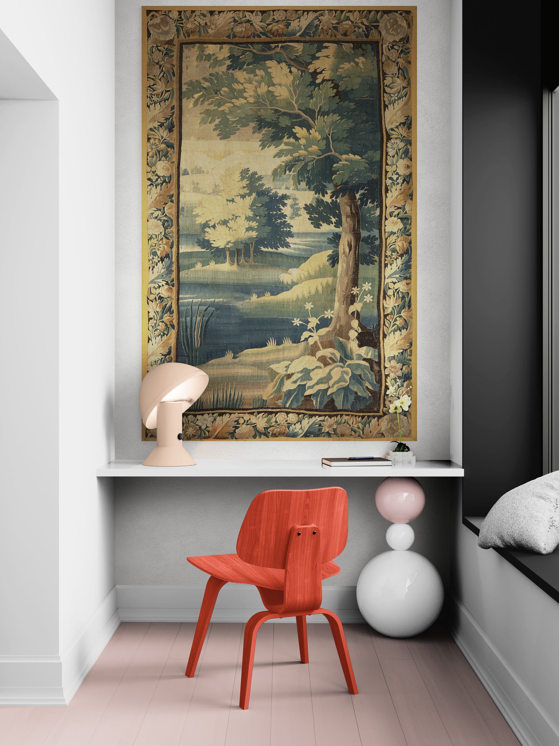 A large vintage-style landscape tapestry with a golden floral border hangs on a white wall above a white desk. In front of the desk is a bright orange-red molded chair, with a beige sculptural table lamp on the left and a white and pink stacked vase on the right.