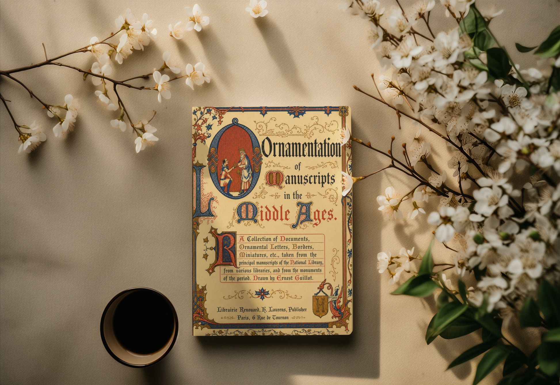 A vintage-style book titled "Ornamentation of Manuscripts in the Middle Ages" lies on a beige surface, surrounded by white blossoms and a small dark cup. The cover has ornate medieval-style illustration and lettering in cream, blue, red, and brown tones.