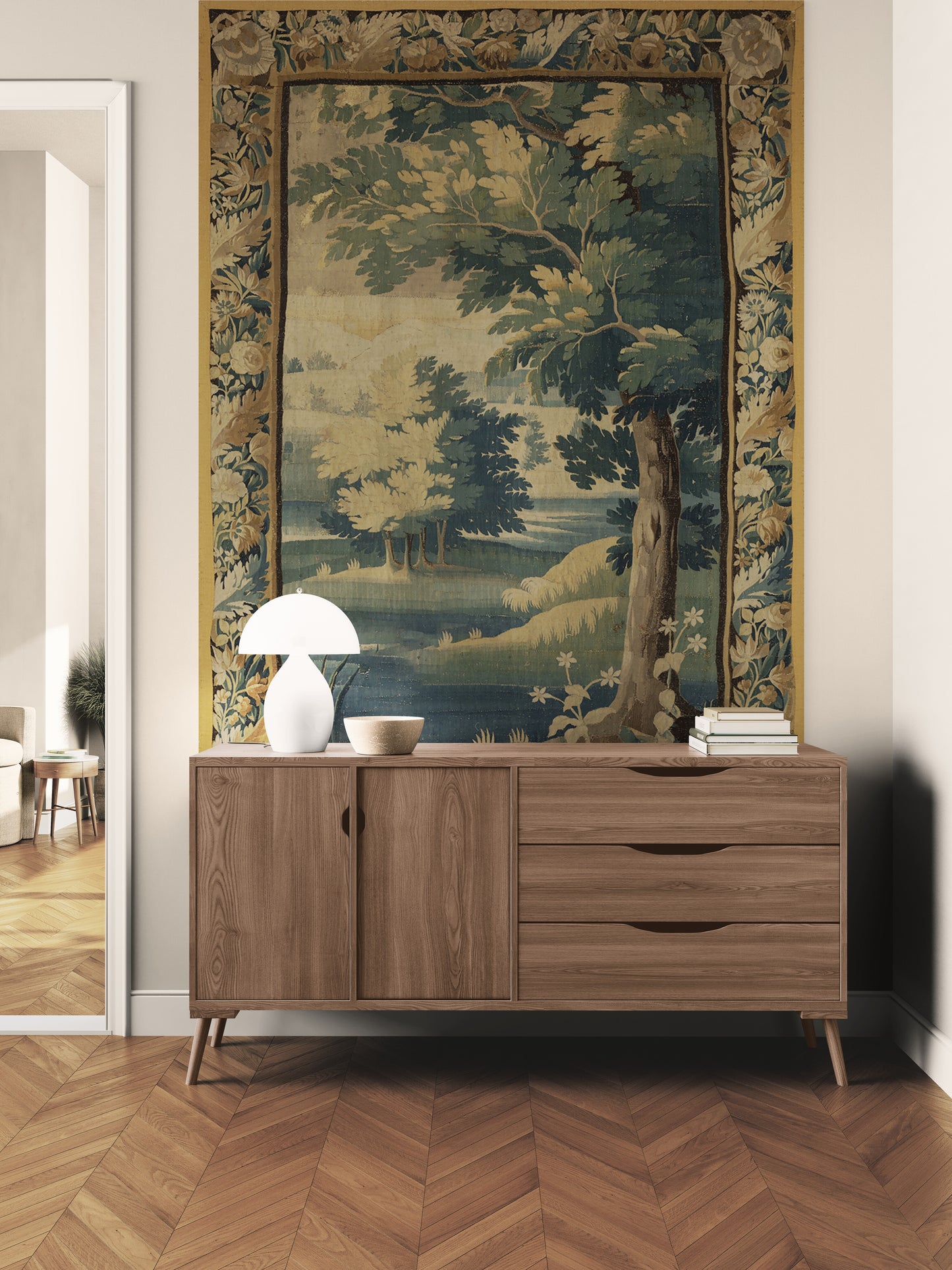 A large tapestry featuring a classical landscape scene with trees and water, styled in a room above a wooden sideboard with a white table lamp and books.