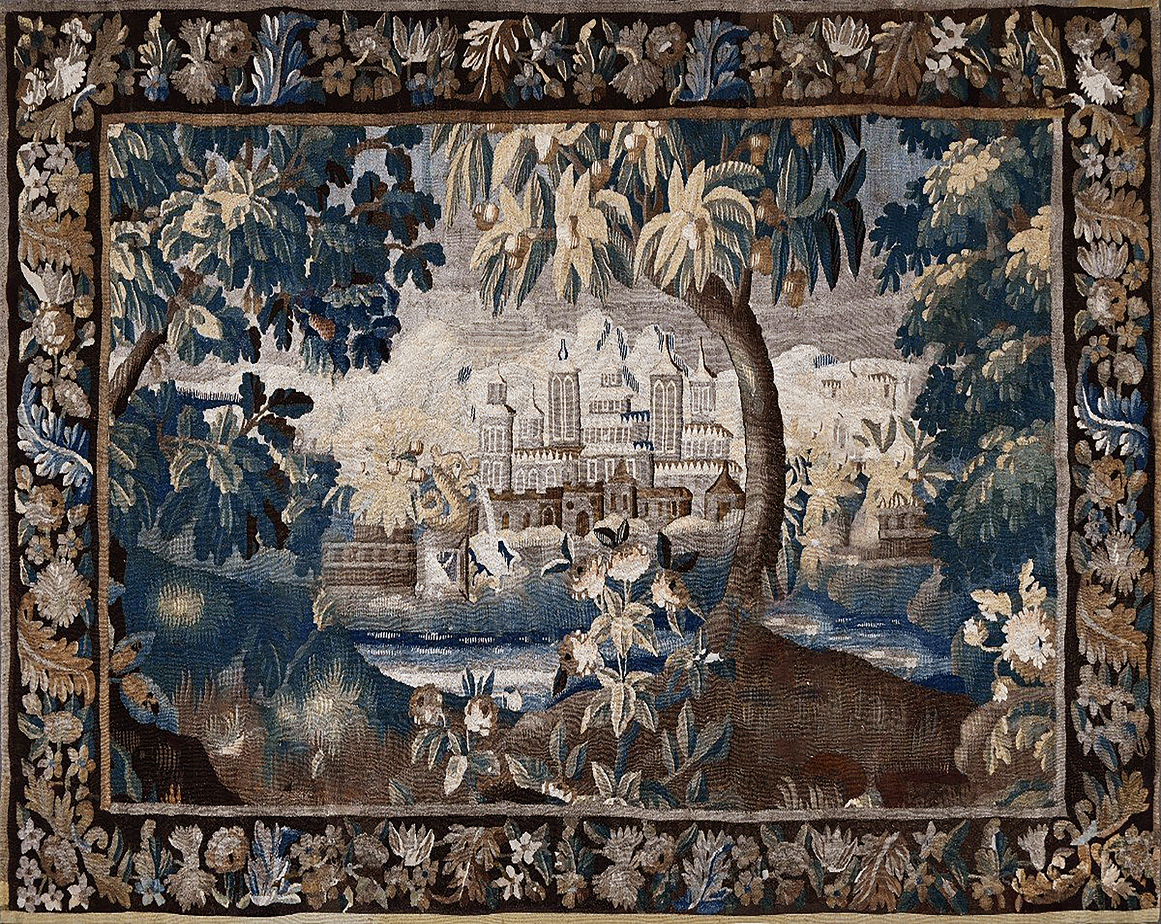 An ornate rectangular tapestry featuring a detailed landscape scene with a grand chateau, trees, water, and figures in the foreground, surrounded by a decorative floral border.