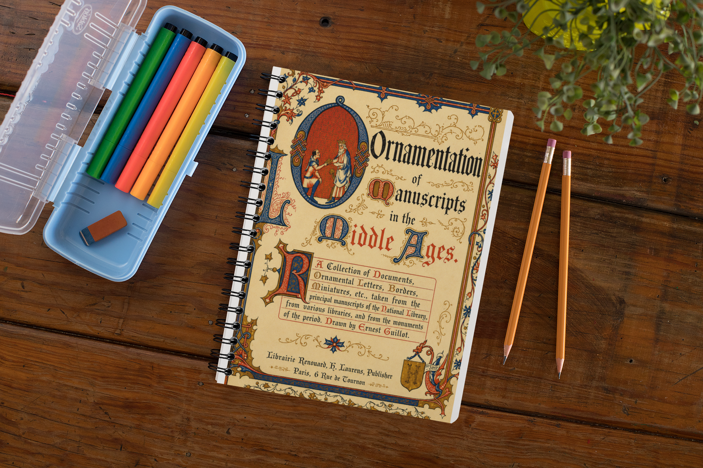 A vintage-style hardcover notebook titled "Ornamentation of Manuscripts in the Middle Ages" lies on a wooden tabletop, surrounded by a blue pencil case with colored pencils, two wooden pencils, and a small plant.