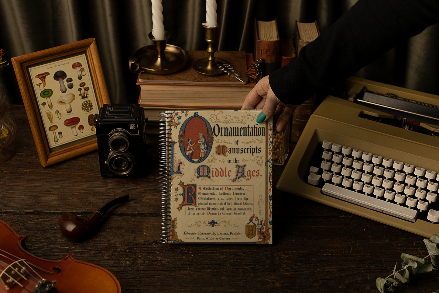 A hand holds a vintage-style book titled 'Ornamentation of Manuscripts in the Middle Ages' on a dark wooden desk surrounded by props including a typewriter, camera, framed mushroom illustration, candles, and a violin.