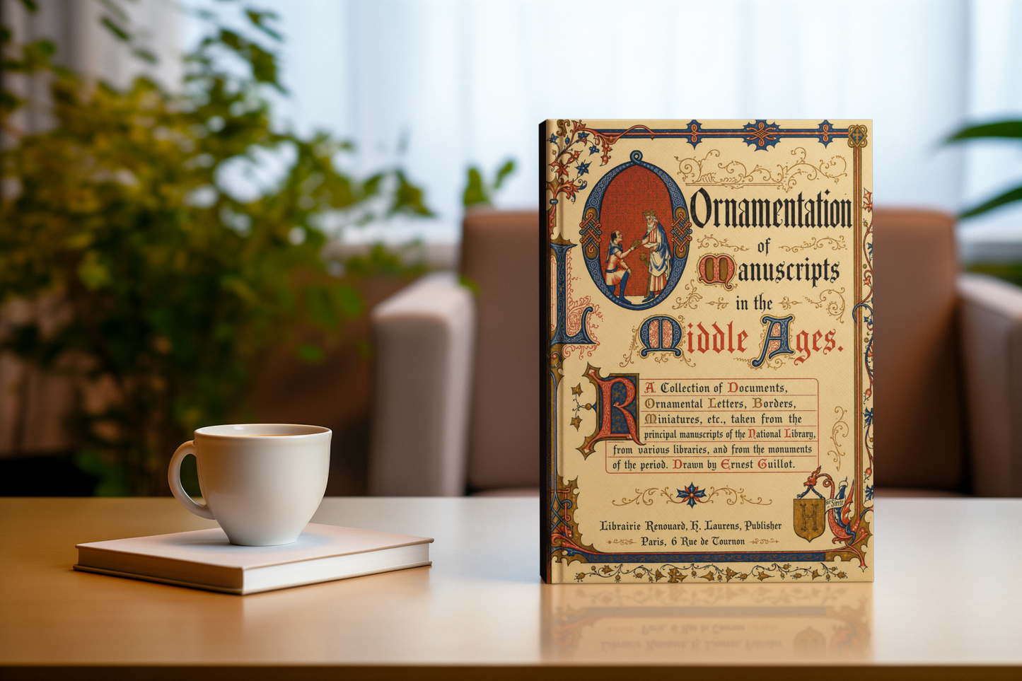A vintage-style book titled "Ornamentation of Manuscripts in the Middle Ages" stands upright on a tabletop beside a white ceramic cup and a small notebook, with a softly blurred indoor background and greenery.