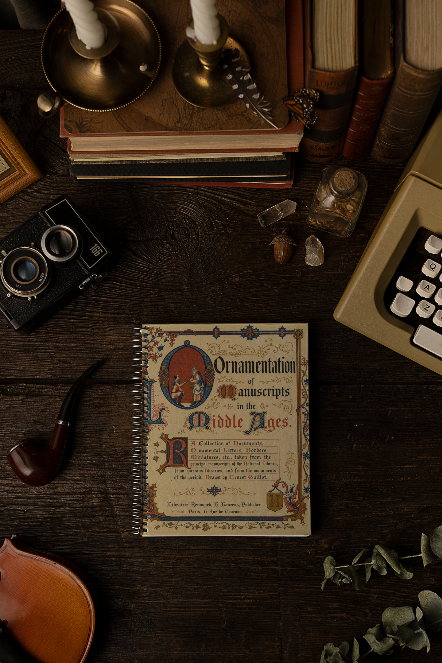 A vintage-style spiral-bound book titled 'Ornamentation of Manuscripts in the Middle Ages' lies on a dark wooden tabletop surrounded by props including a camera, typewriter, books, candlesticks, a pipe, and a violin.